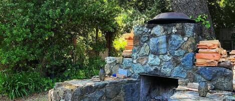 Un espace barbecue en pierre, avec des chaises en bois et un grand arbre en arrière-plan.