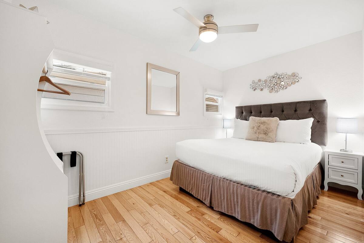 A bedroom with a large bed, a ceiling fan, a bedside table with a lamp, and a window with blinds.