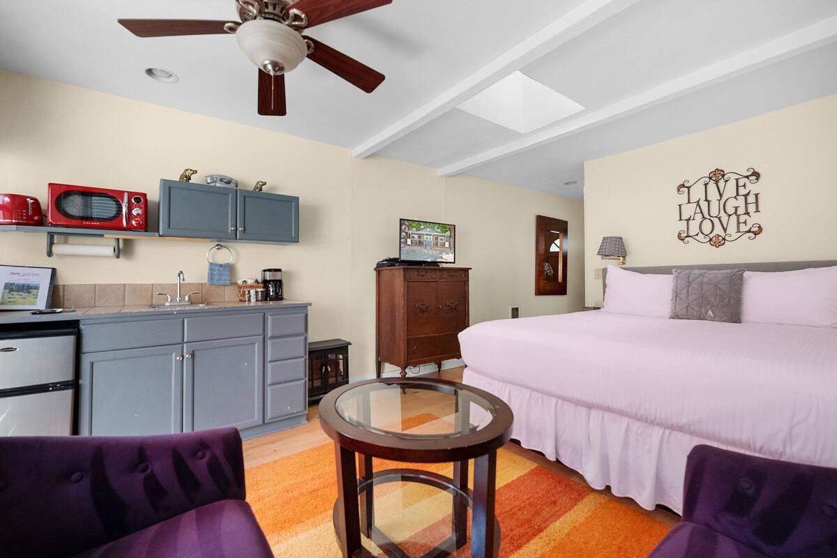 A compact bedroom with a bed, a small table, a ceiling fan, and a kitchenette with a microwave.