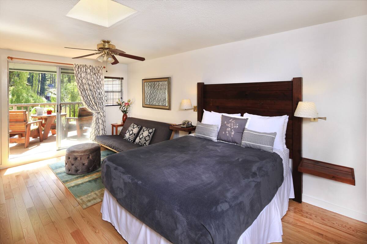 A bedroom with a large bed, a ceiling fan, a nightstand, a lamp, and a view of the outdoors through a sliding glass door.