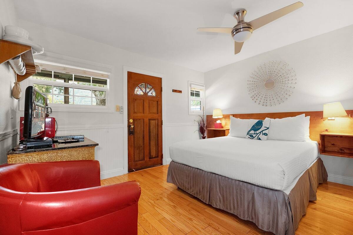 A bedroom with a bed, a red chair, a wooden desk with a lamp, a ceiling fan, and a decorative wall piece.