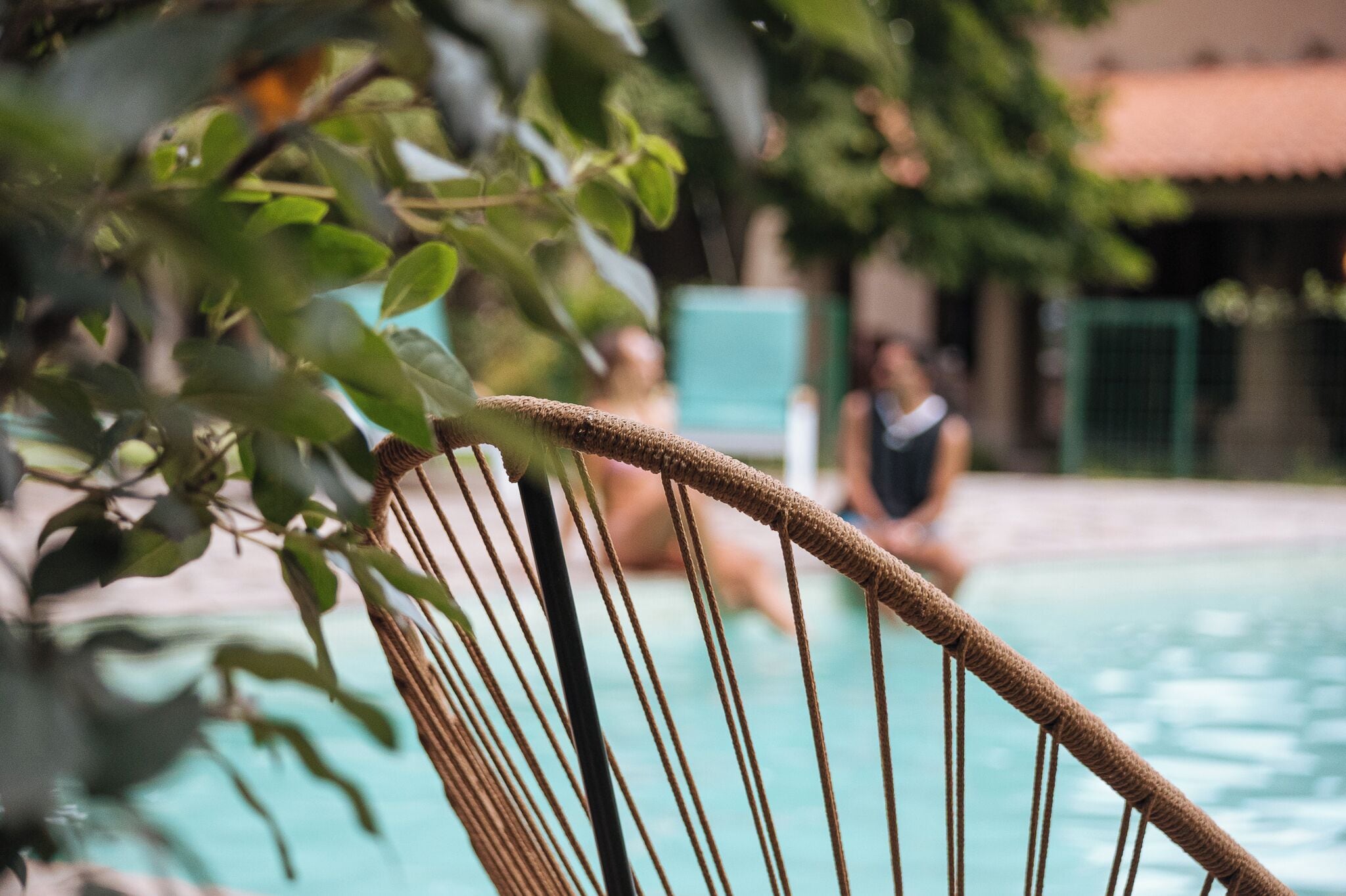 Una silla de mimbre junto a una piscina, con un fondo borroso de un edificio y vegetación.