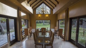 Un vaste espace dédié à la salle à manger, avec une table en verre entourée de chaises, de grandes fenêtres donnant sur un jardin et un lustre au-dessus.