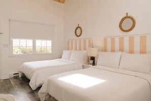 A hotel room with two beds, a window, a bedside table with a lamp, and a wall-mounted mirror.