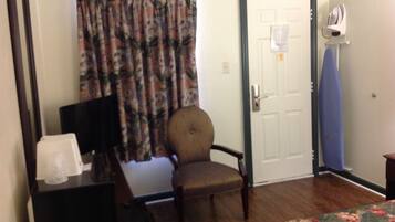 A room with a wooden floor, a floral-patterned bed, a chair, a desk with a lamp, and a door with a paper on it.