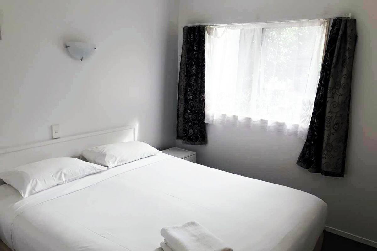 A single-bed hotel room with a neatly made bed, a bedside table, a wall-mounted light, and a window with curtains.