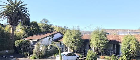 Un estacionamiento con un coche blanco, rodeado de follaje y un edificio con tejado de tejas.
