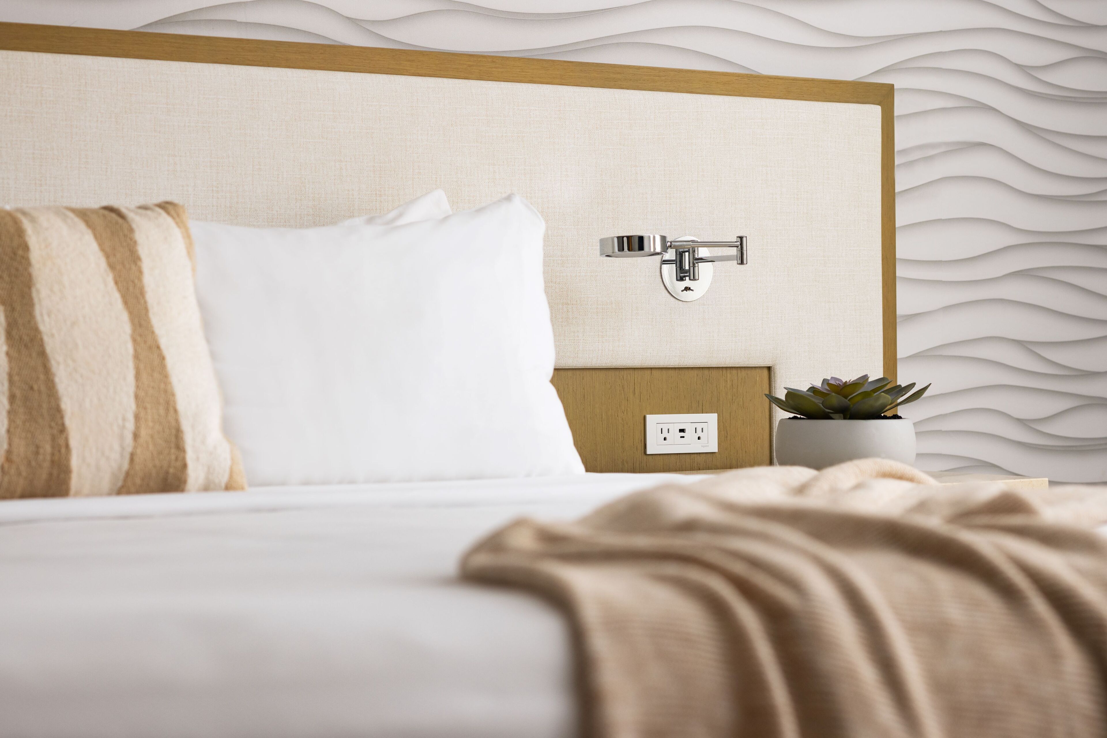 A neatly made bed with white and striped pillows, a beige blanket, a wall-mounted power outlet, and a small potted plant.