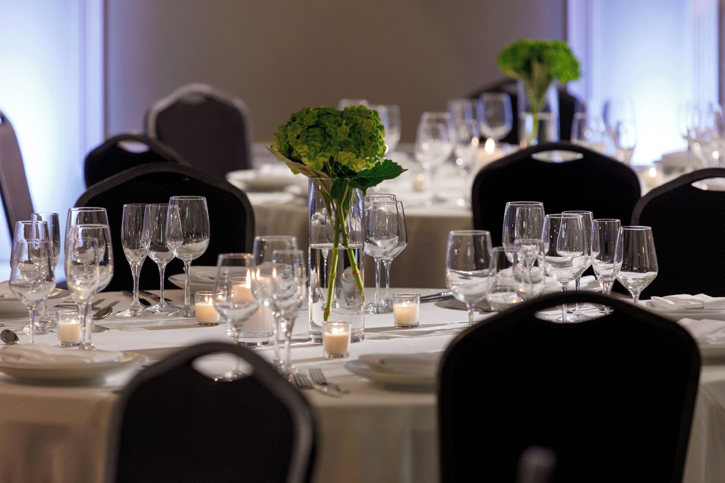 Una mesa de comedor formal dispuesta con copas de vino, platos y cubiertos, junto a velas y un florero con follaje verde.