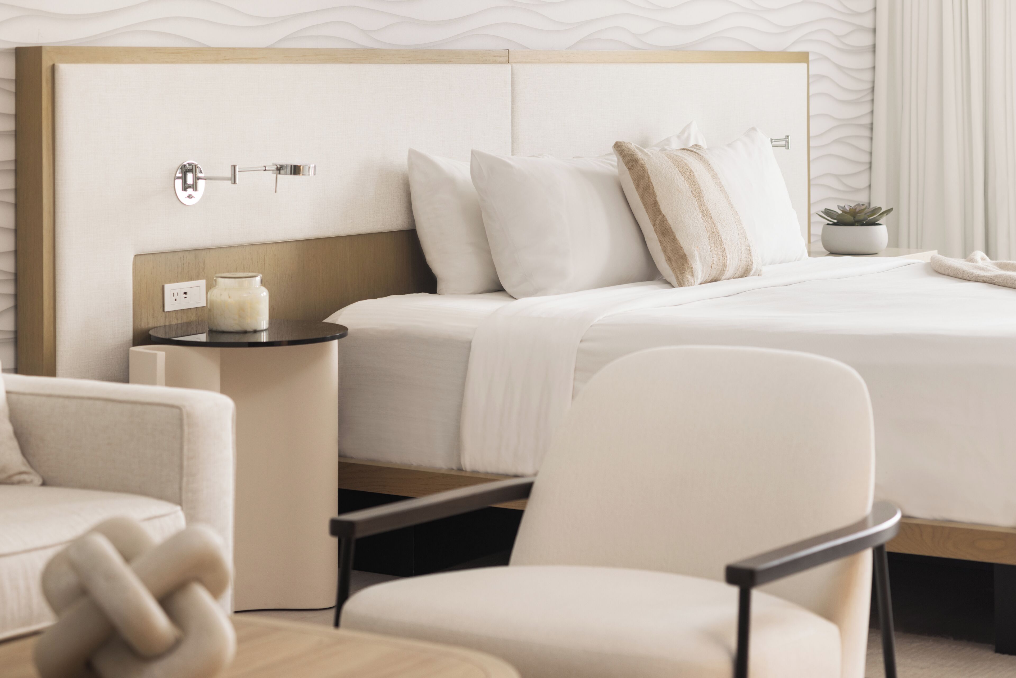Una habitación de hotel moderna con una cama grande, una mesita de noche con lámpara, una silla y una pequeña planta en maceta.
