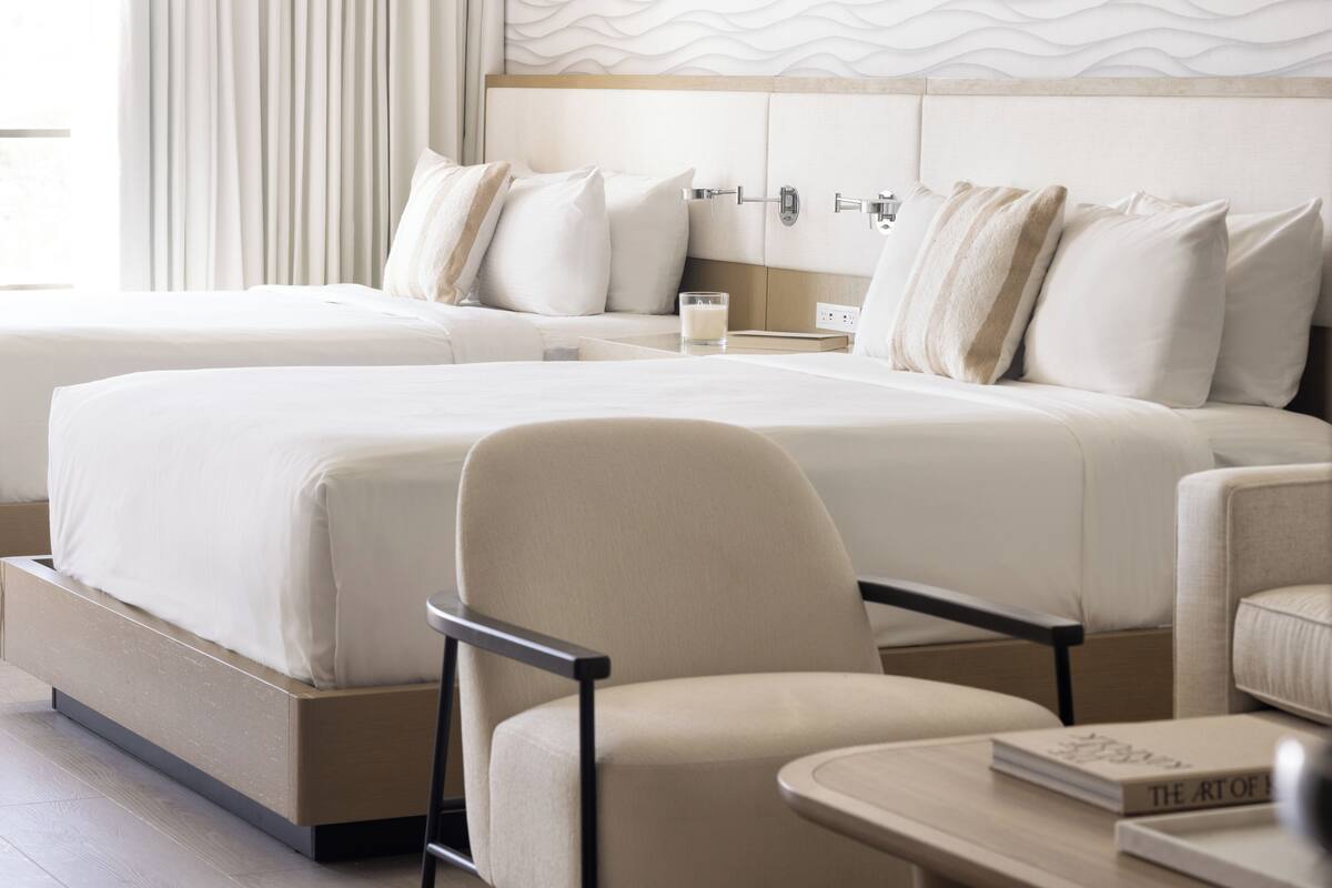 Une chambre d’hôtel avec deux lits, une chaise, une table de chevet et un livre intitulé « L’art de… ».
