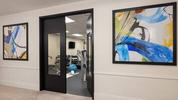 Un pasillo con tres cuadros abstractos enmarcados, una puerta que da a un área de gimnasio y una pared blanca con piso texturizado.