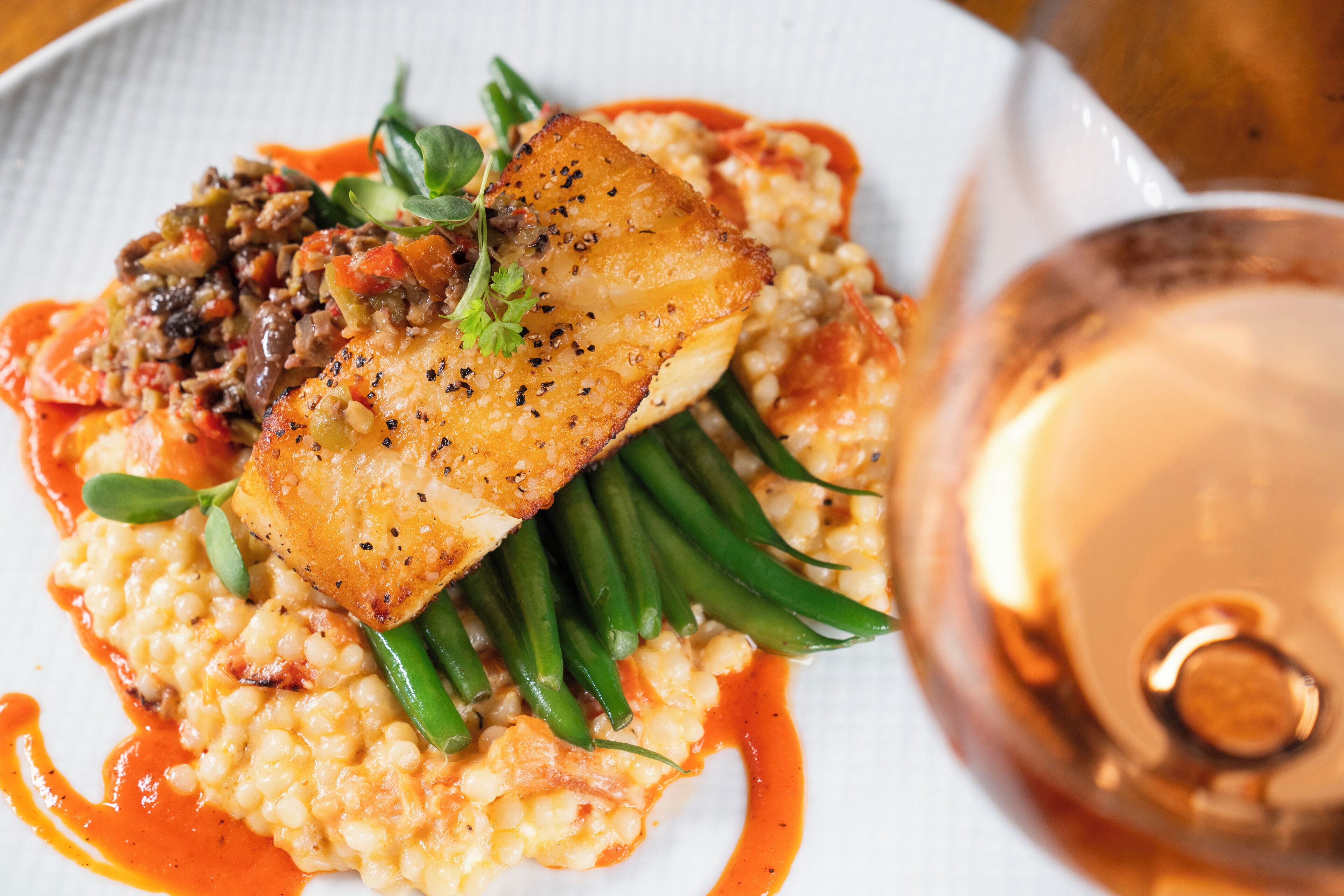 Une assiette composée d’un filet de poisson, de haricots verts et d’une sauce cr émeuse, accompagnée d’un verre de vin.