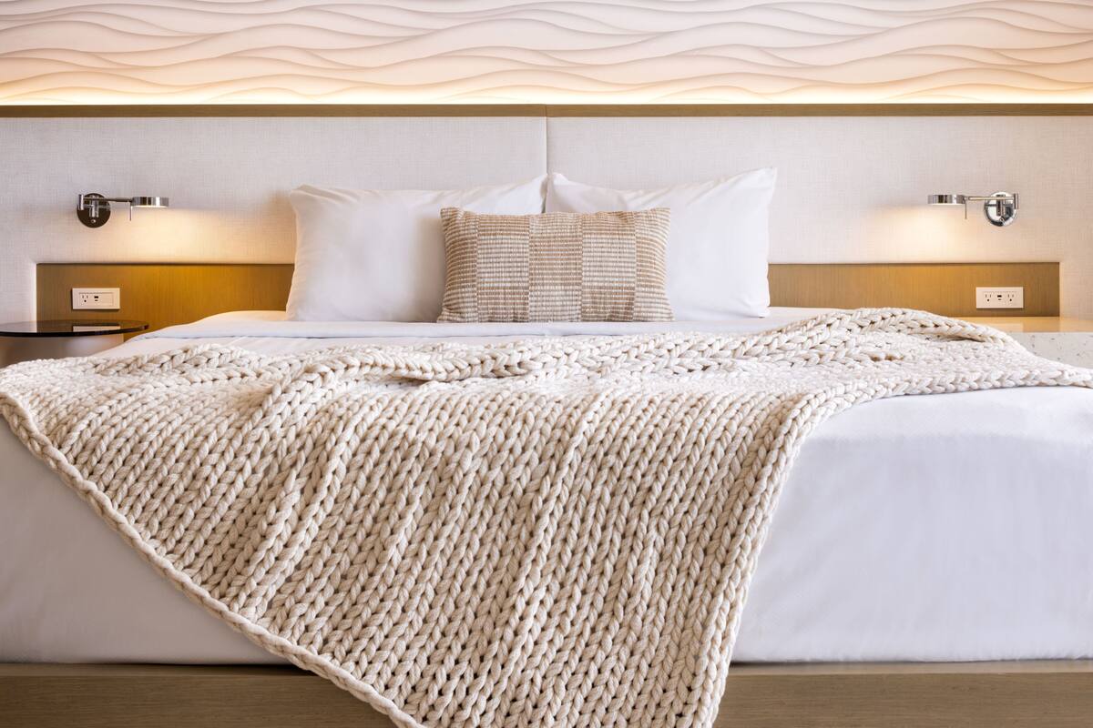Un lit bien fait, recouvert d’une couverture tricotée, une tête de lit au motif texturé et des lampes de chevet de chaque côté.