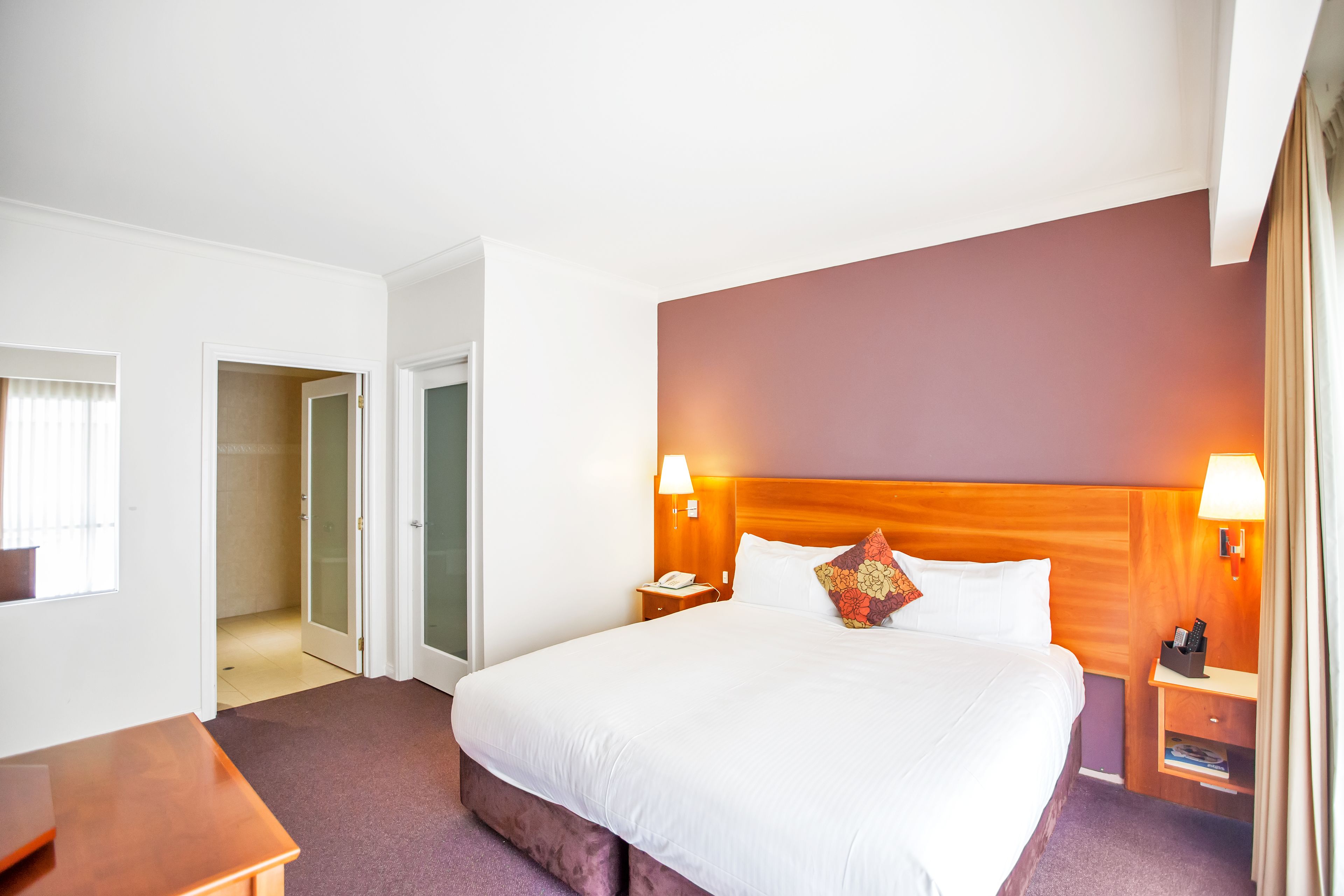 A hotel room with a large bed, two bedside lamps, a wooden headboard, a nightstand with a phone, and a small desk.
