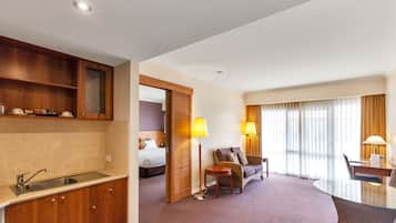 A hotel room with a kitchenette, a bed, a sitting area with a sofa and armchairs, and a dining table with lamps.
