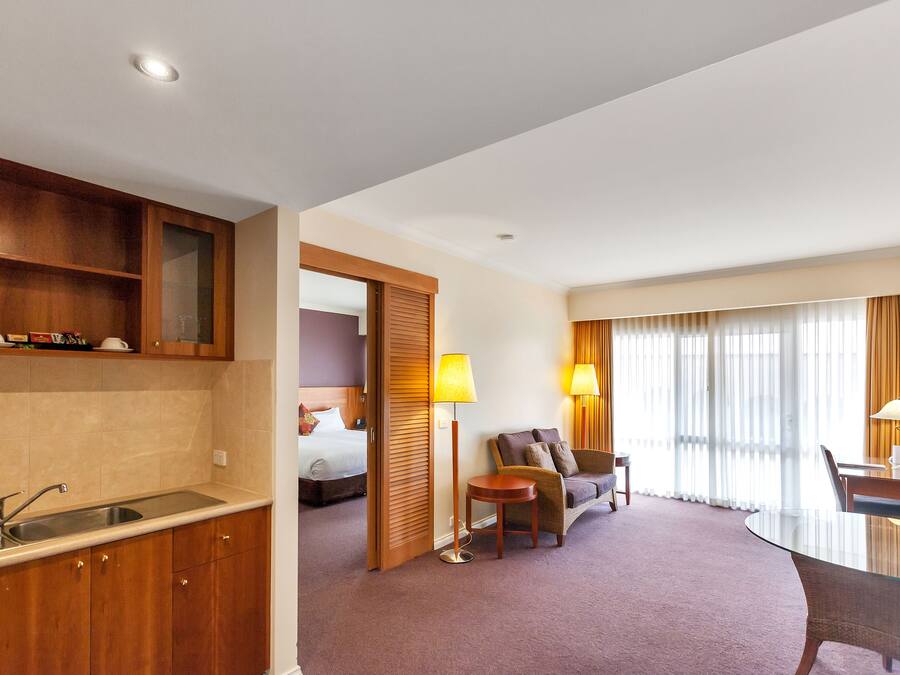 A hotel room with a kitchenette, a bed, a sitting area with a sofa and armchairs, and a dining table with lamps.