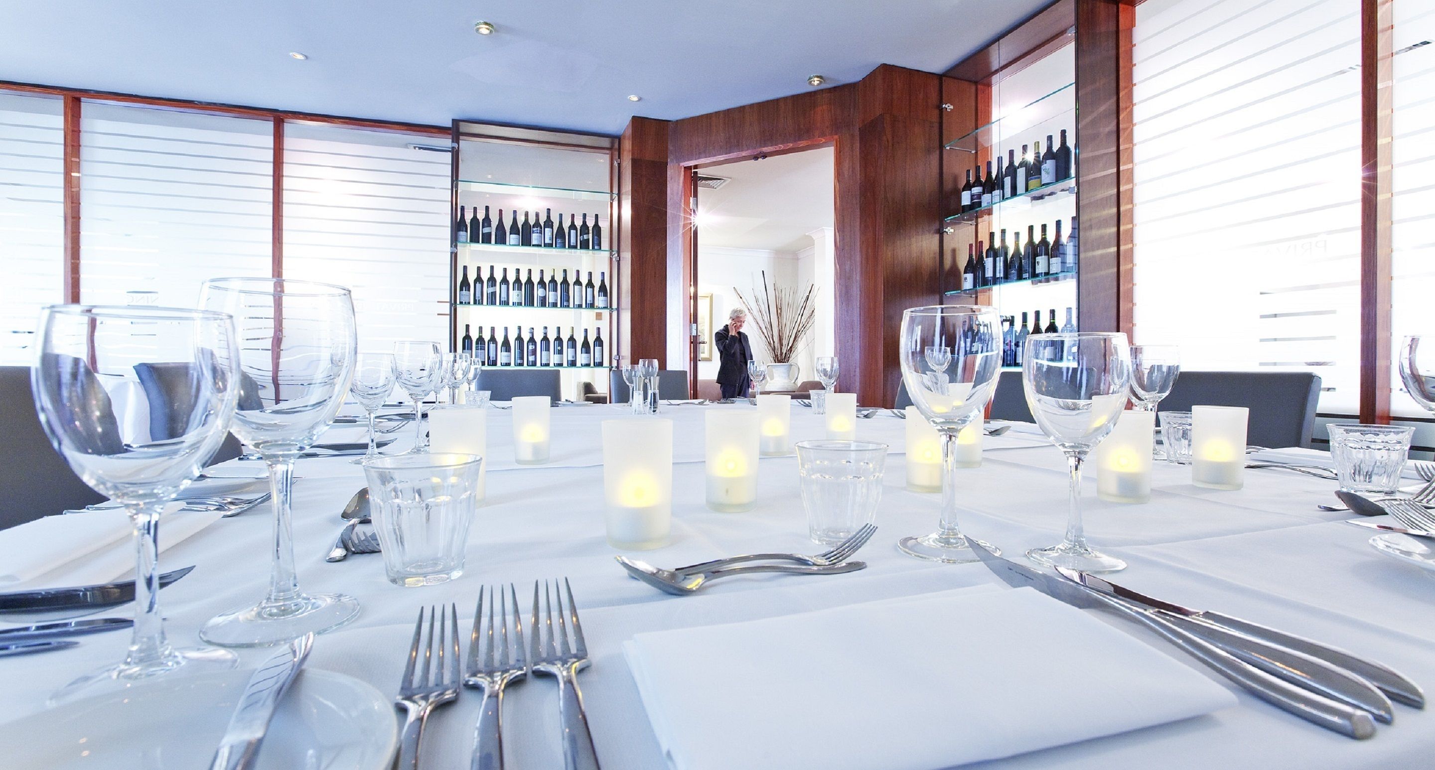 A well-lit dining area with a long table set for a meal, featuring wine glasses, cutlery, and candles.