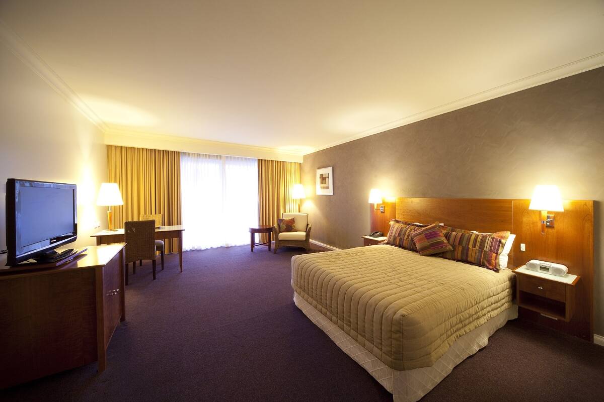 A hotel room with a large bed, a desk with a television, two chairs, a small table, and a nightstand with a lamp.
