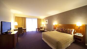 A hotel room with a large bed, a desk with a television, two chairs, a small table, and a nightstand with a lamp.