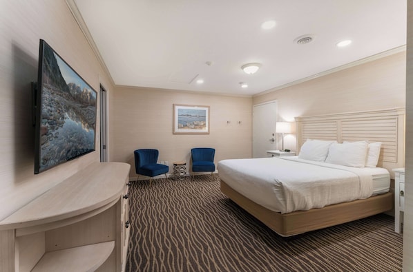 Habitación de hotel con una cama grande, dos sillas azules, un escritorio con televisor de pantalla plana, un cuadro en la pared y una alfombra estampada.