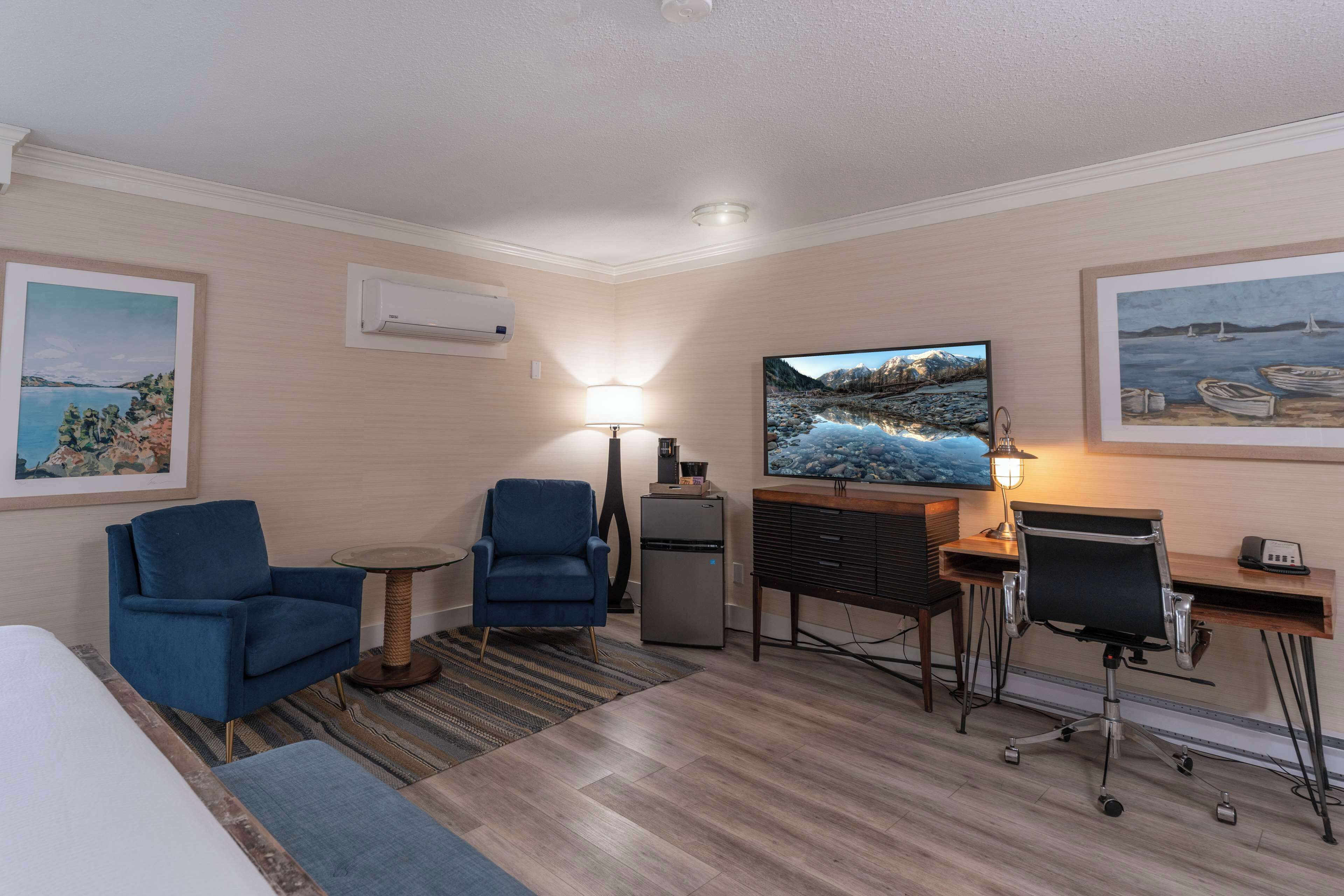 A hotel room with a bed, two blue armchairs, a desk with a computer, a TV, and a refrigerator.