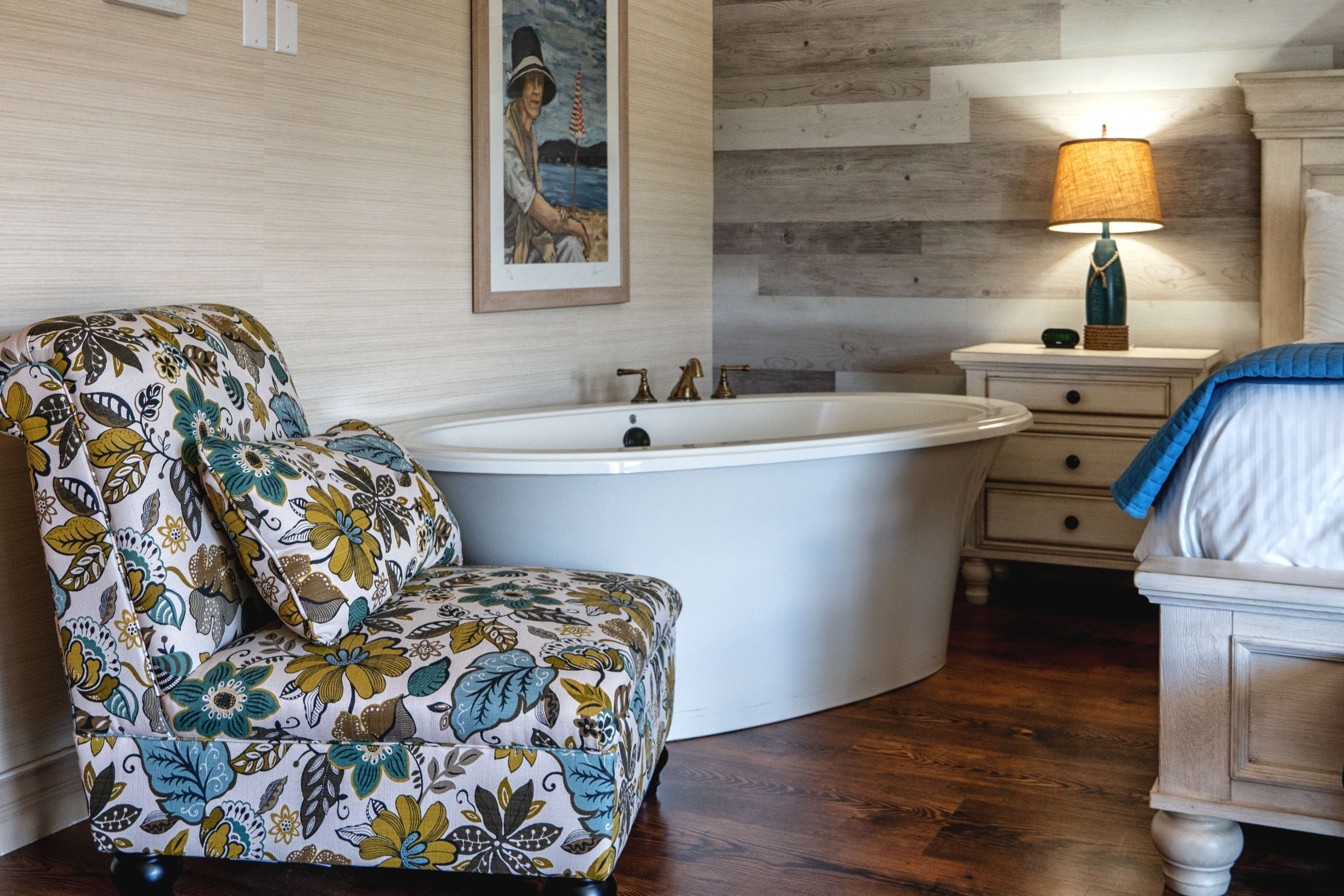 A bathroom with a bathtub, a floral-patterned chair, a wooden floor, and a nightstand with a lamp.
