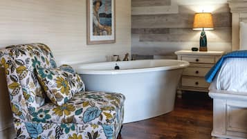 A bathroom with a bathtub, a floral-patterned chair, a wooden floor, and a nightstand with a lamp.
