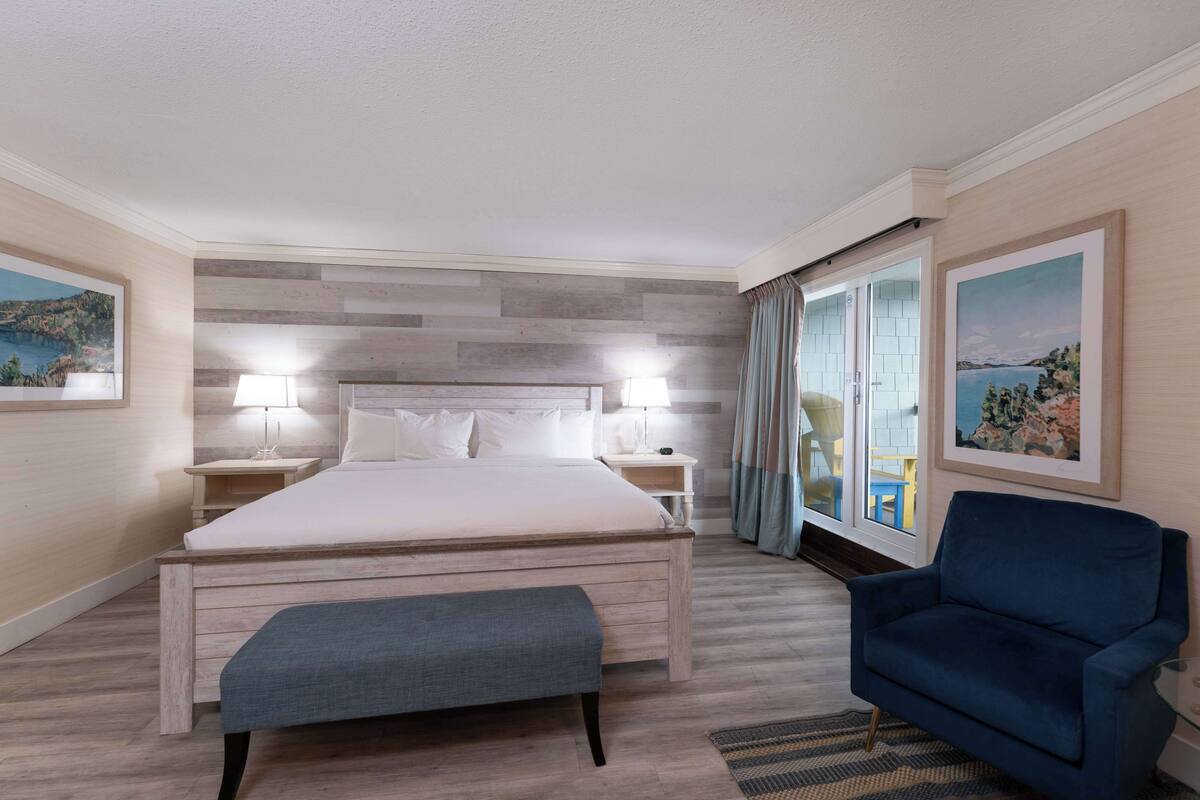 A bedroom with a bed, bedside tables, a bench, a blue armchair, and a view of the outdoors.