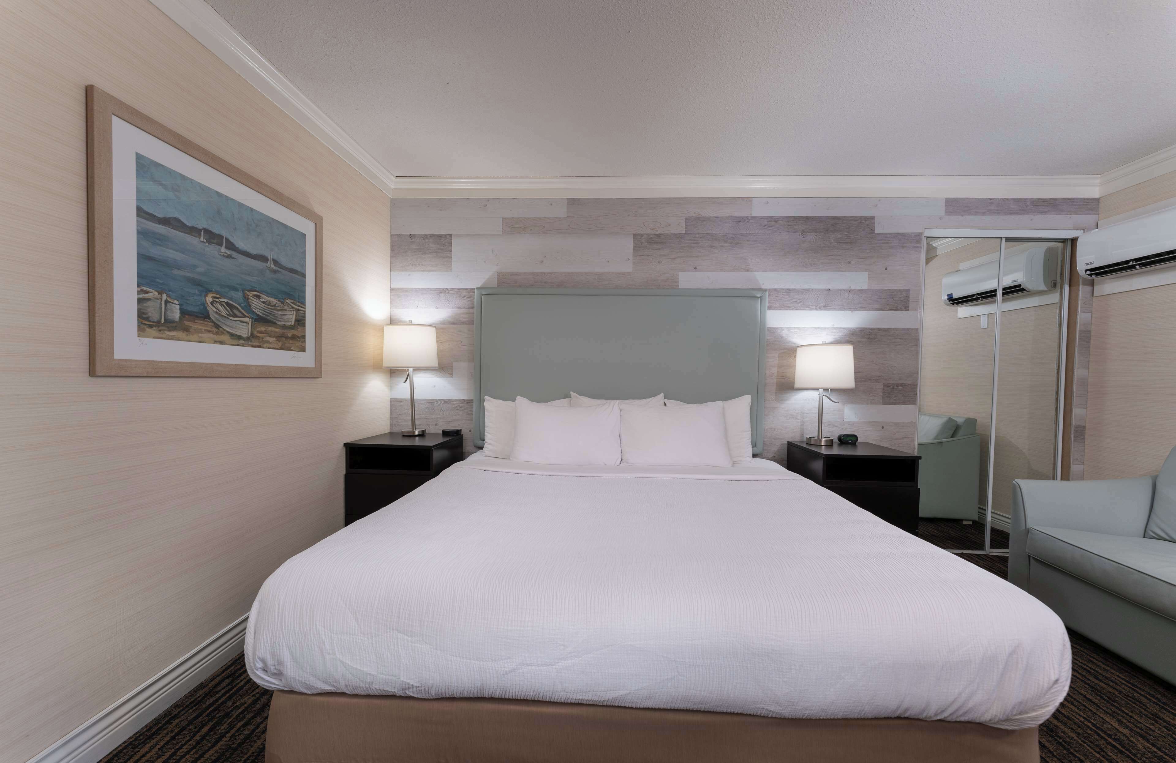 A hotel room with a large bed, two bedside tables with lamps, a grey headboard, a framed painting on the wall, and a walk-in closet with a mirror.