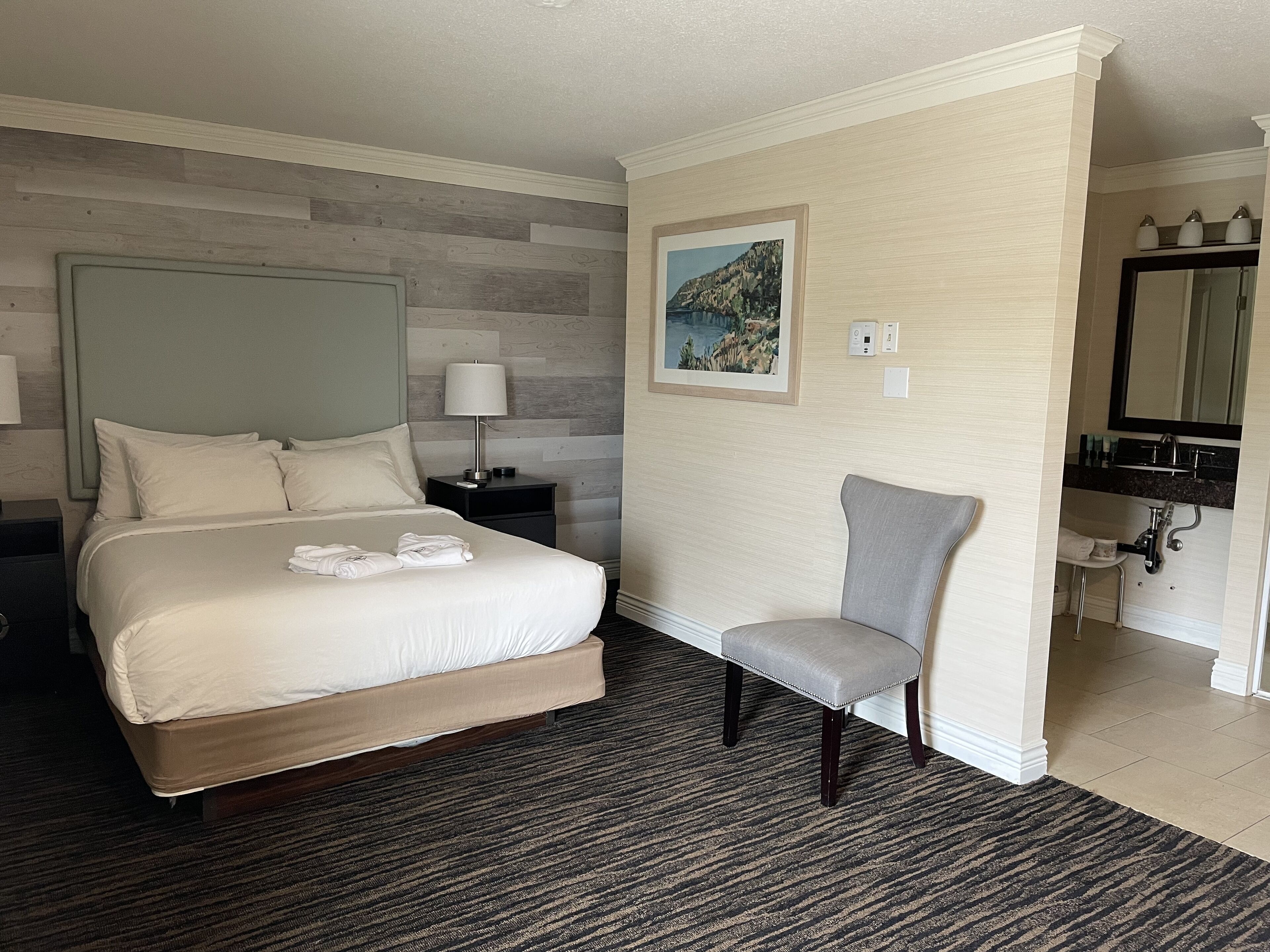Una camera d'albergo con un letto, una sedia, un comodino con una lampada, un quadro appeso al muro e un bagno visibile tramite una porta aperta.