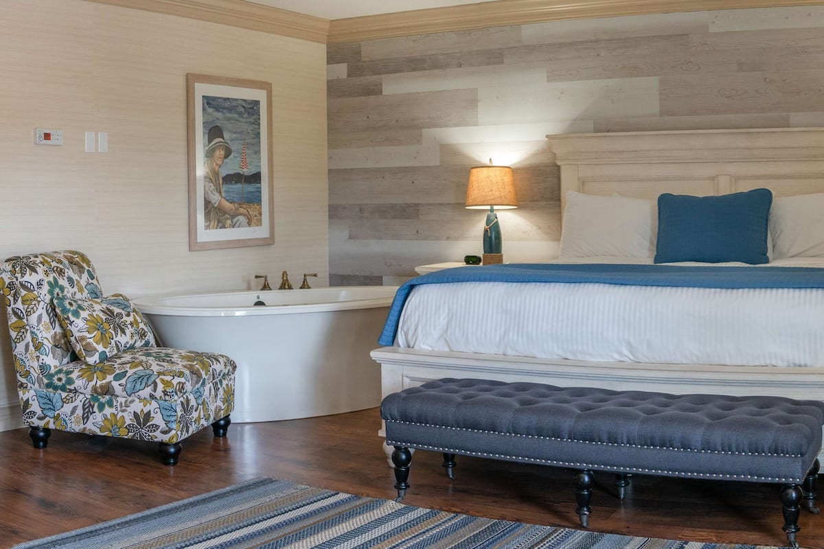 A bedroom with a large bed, two bedside tables with lamps, a chair, a bench, a bathtub, and a framed picture on the wall.