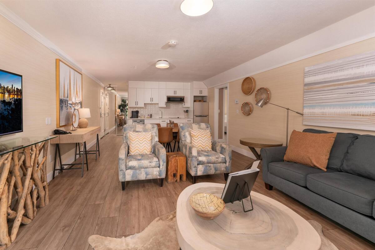 A modern living room with a grey sofa, a coffee table, and a TV. There is a kitchen in the background with white cabinets and a dining area with a table and chairs.