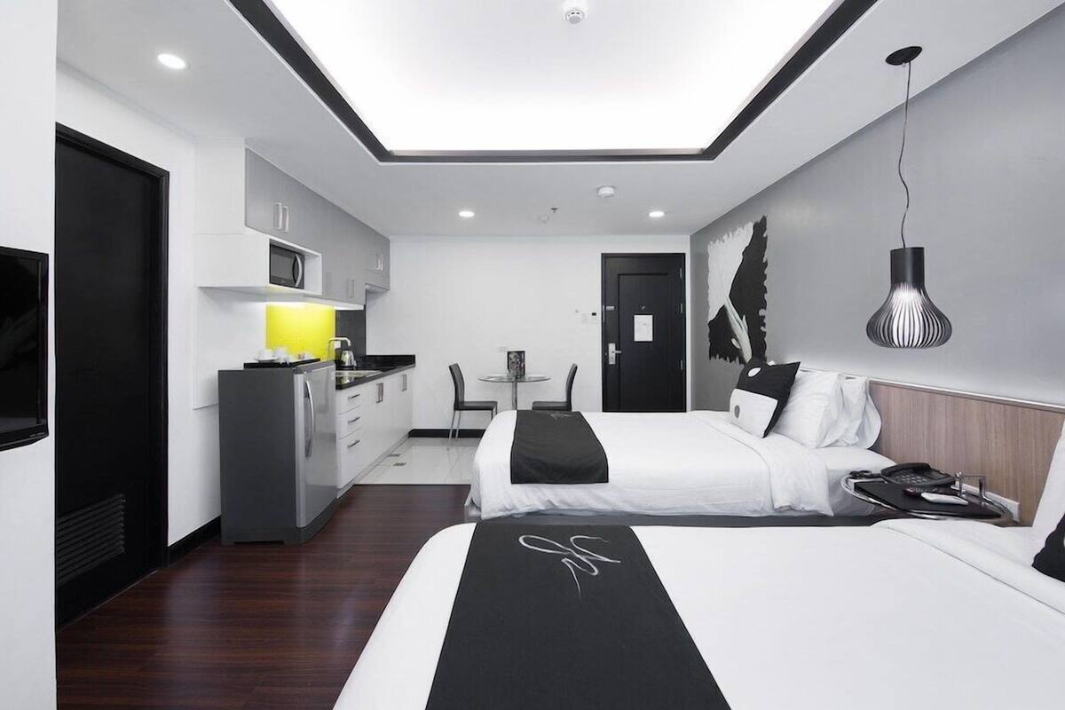 A modern hotel room with two beds, a kitchenette, a dining area, and a large window.