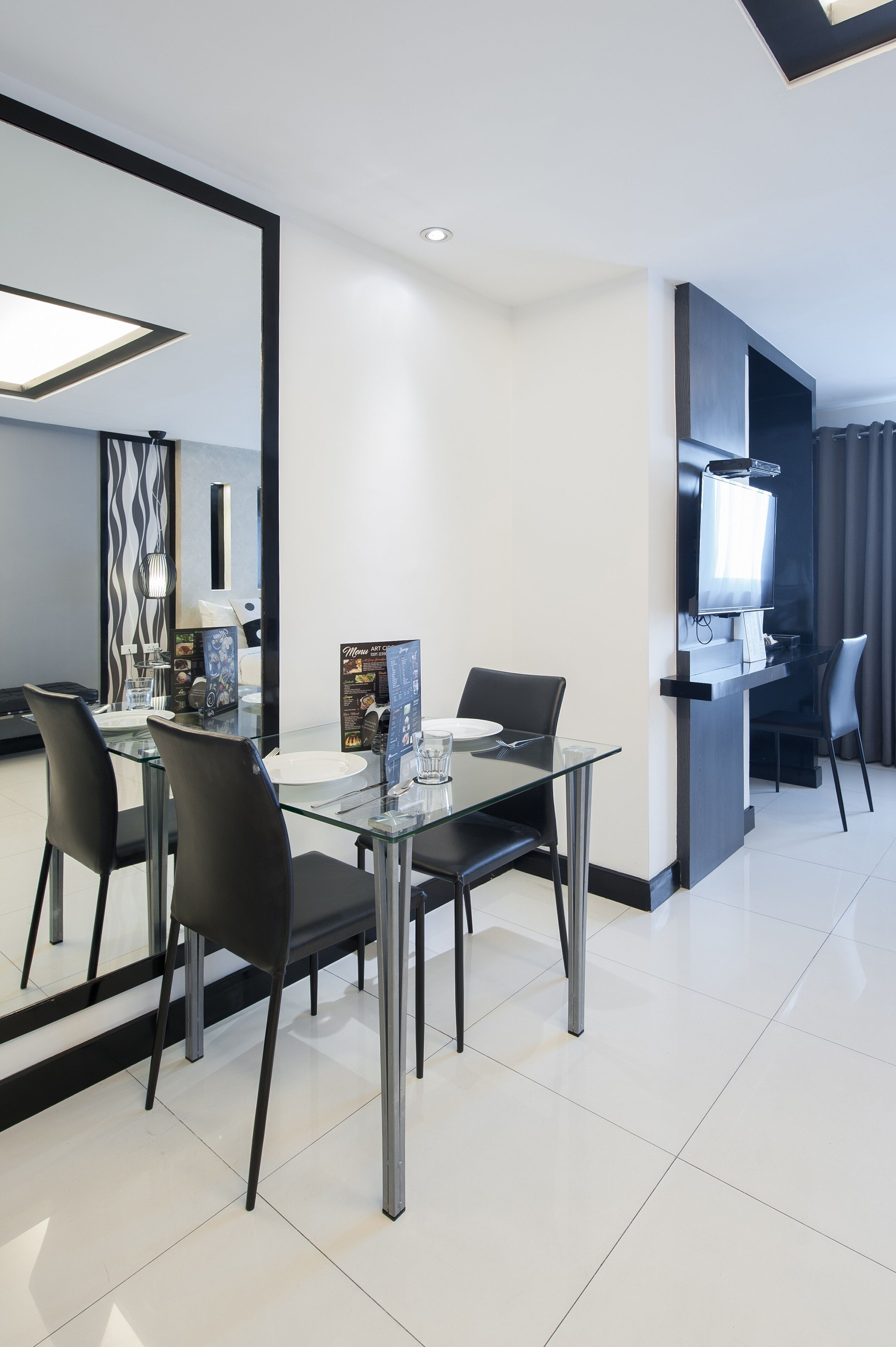 Une chambre d’hôtel moderne avec une table à manger en verre, des chaises noires, un téléviseur à écran plat et un grand miroir.