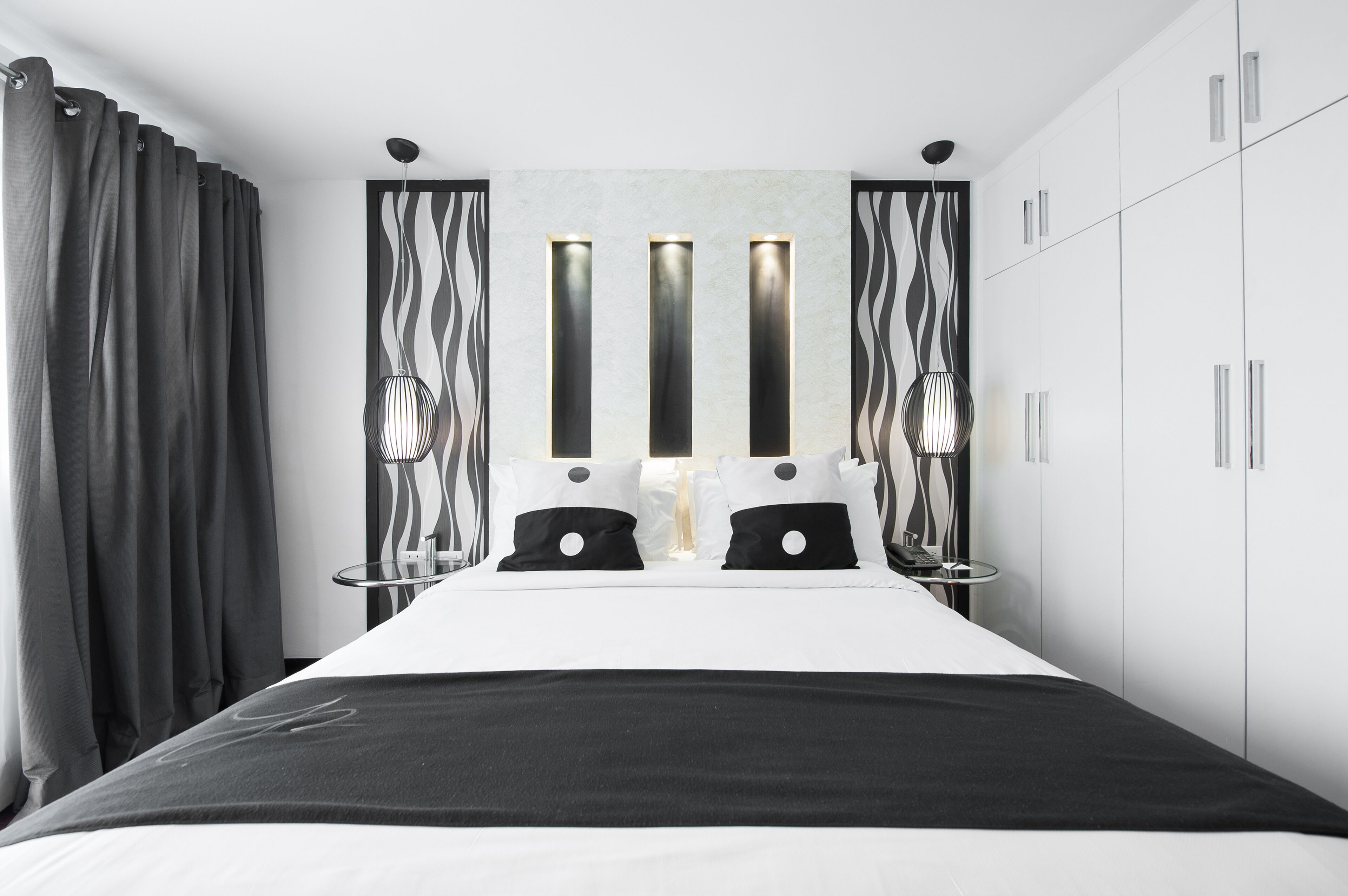 A modern hotel room with a large bed, black and white curtains, a wall-mounted light fixture, and a wardrobe.