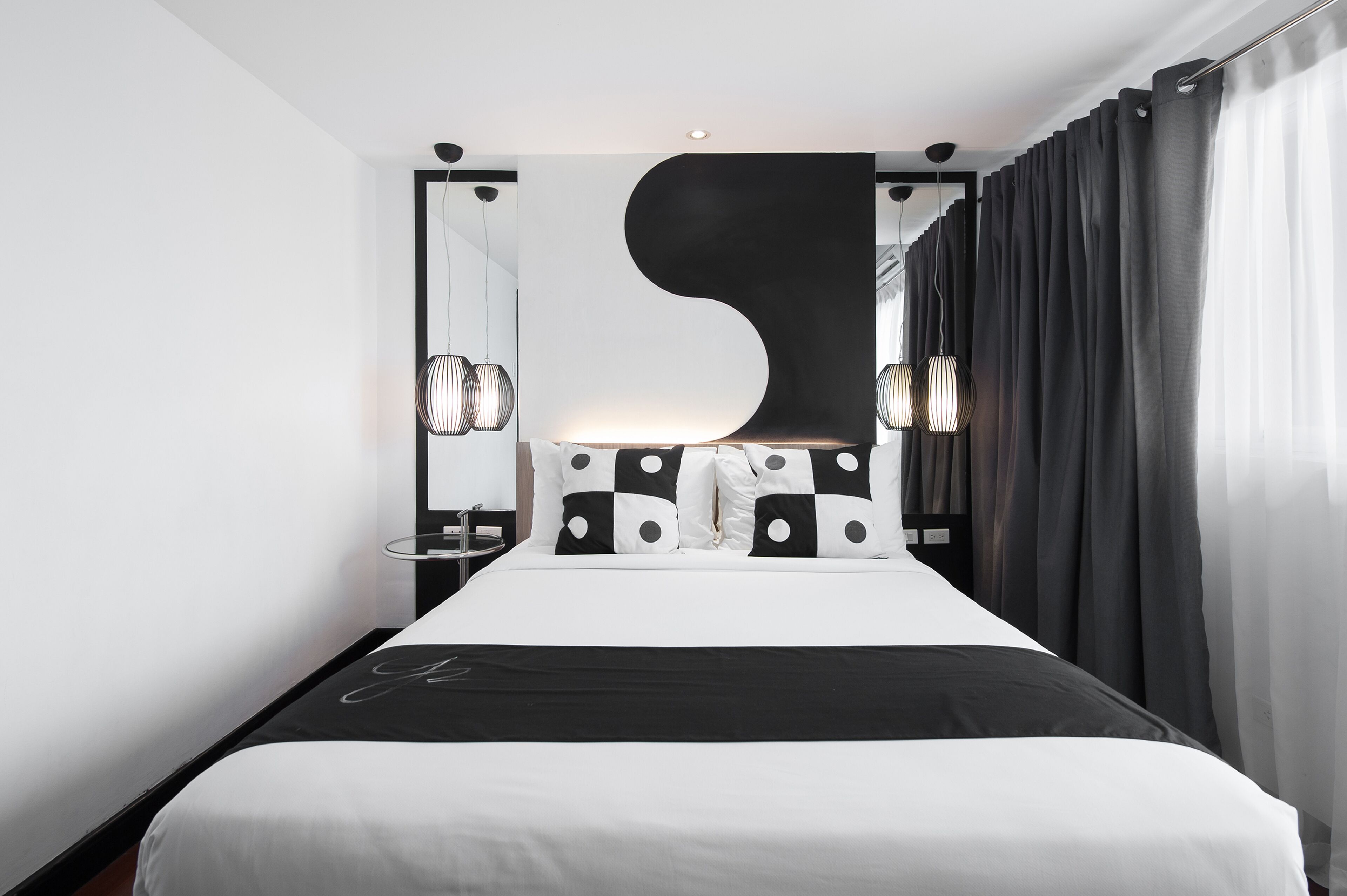 Ein modernes Hotelzimmer mit einem großen Bett, schwarz-weiß gemusterten Kissen, einem einzigartigen schwarzen Wandkunstwerk und einem großen Fenster mit Vorhängen.