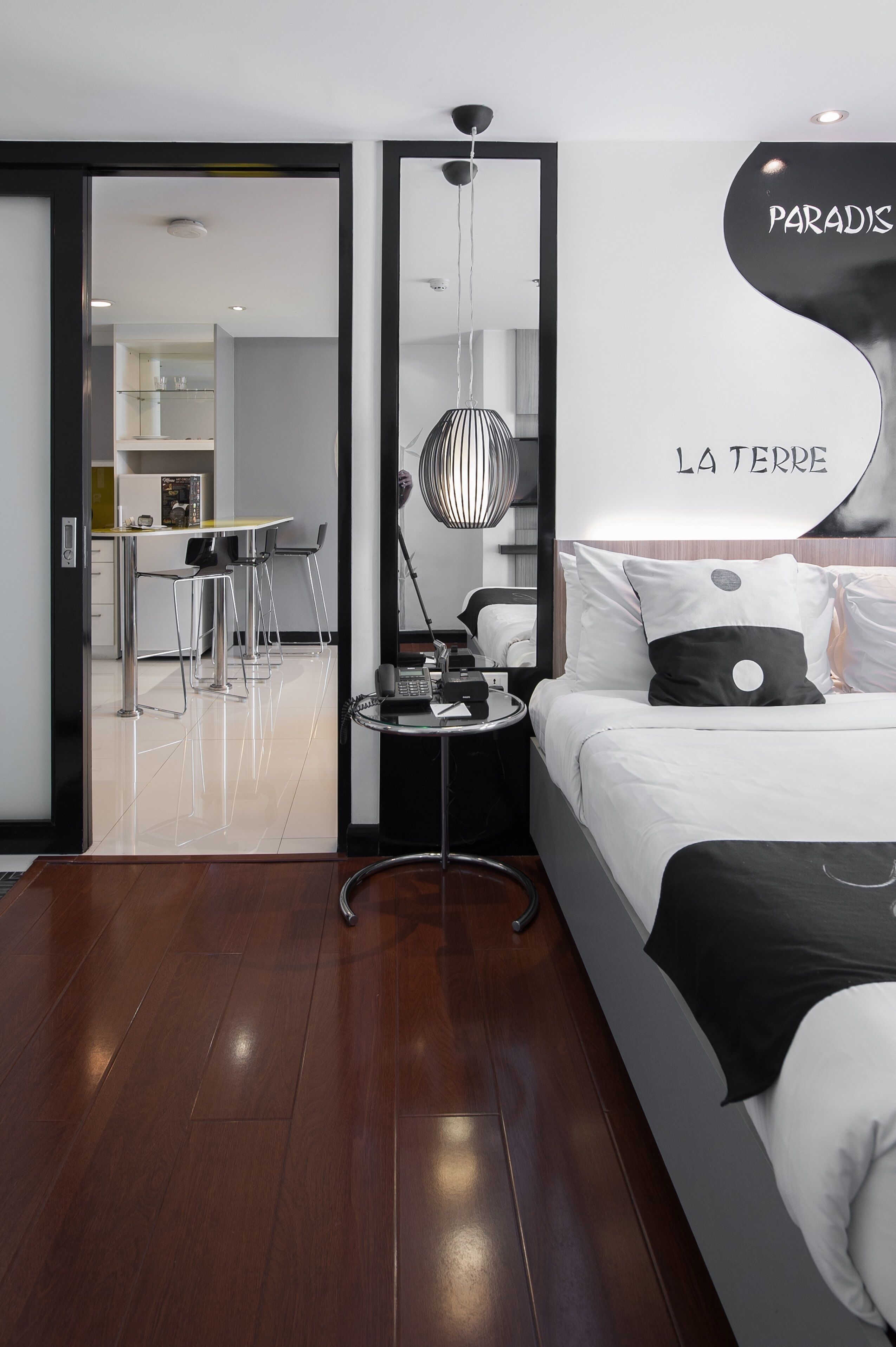 Een moderne hotelkamer met een bed, nachtkastje en een spiegel. Er is een zithoek met stoelen en een tafel zichtbaar door een geopende deur.