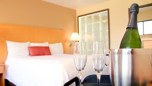 Una camera d'albergo con un letto, un comodino, una lampada, una finestra e un tavolo con una bottiglia di champagne e bicchieri.