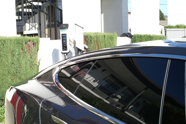 A black car parked near a white building with a staircase and a green hedge.