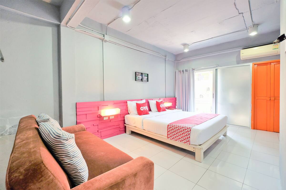 Ein Hotelzimmer mit einem Bett, einer pinkfarbenen Kopfteil, einer roten und weißen Bettdecke, einem gestreiften Kissen, einem weißen Sofa und einem Fenster mit Vorhängen.