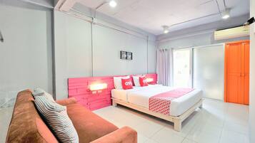 Quarto de hotel com cama, cabeceira rosa, colcha vermelha e branca, travesseiro listrado, sofá branco e janela com cortinas.