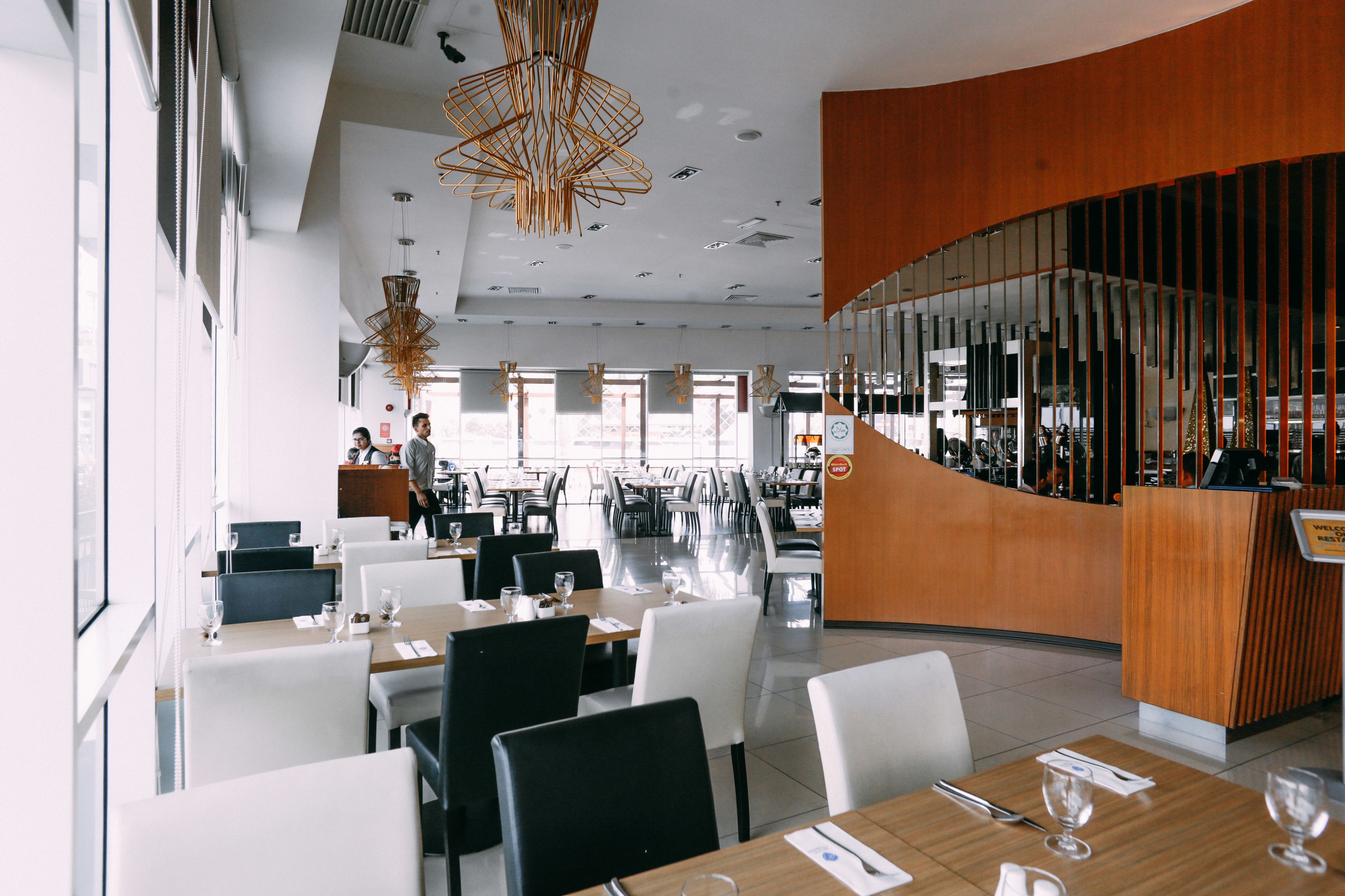 A modern restaurant with a curved wooden reception desk, multiple dining tables with chairs, and hanging pendant lights.