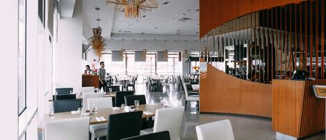 A modern restaurant with a curved wooden reception desk, multiple dining tables with chairs, and hanging pendant lights.