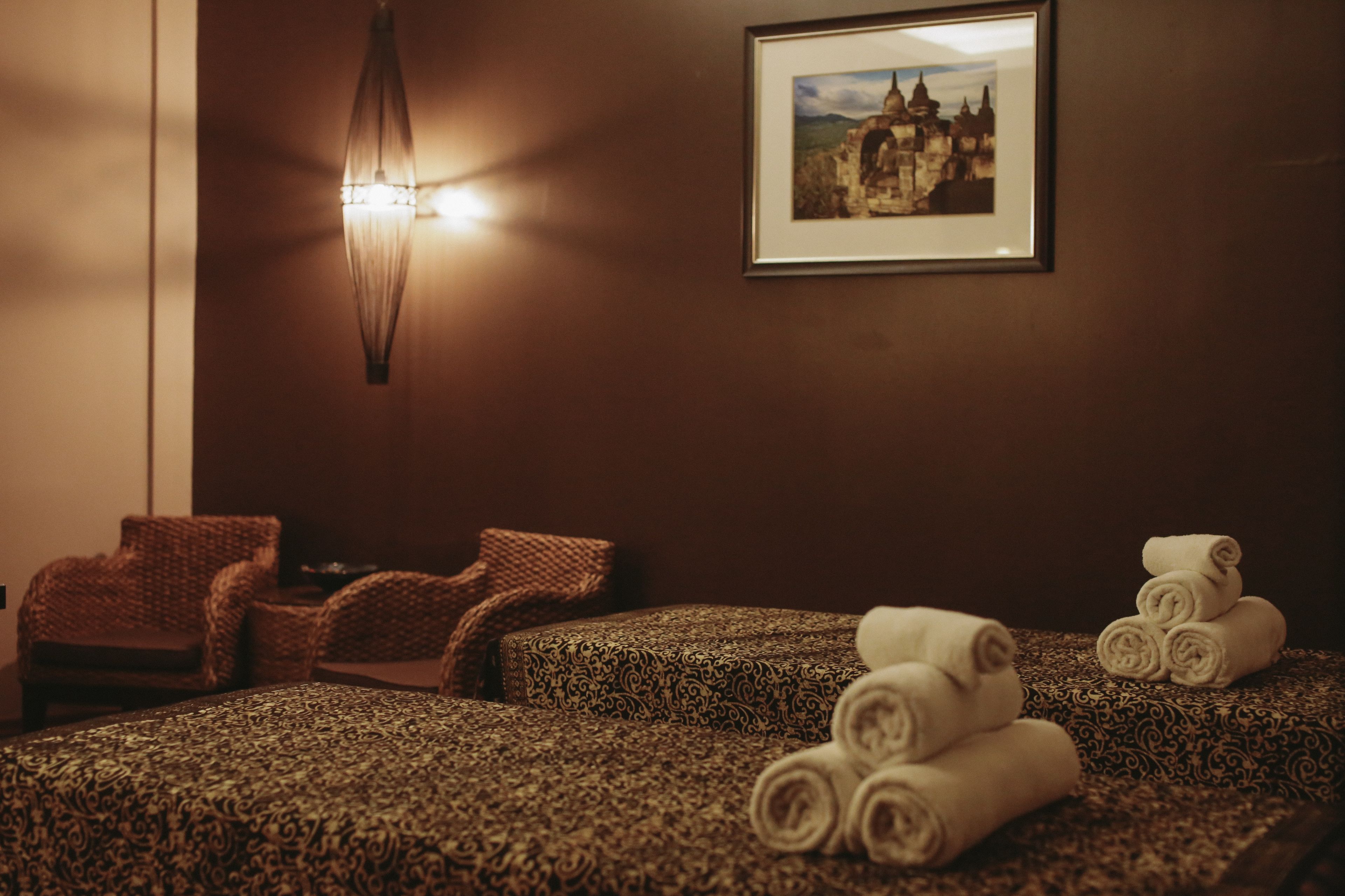 Um quarto de spa com carpete estampado, duas cadeiras de vime, um quadro na parede e toalhas enroladas no chão.