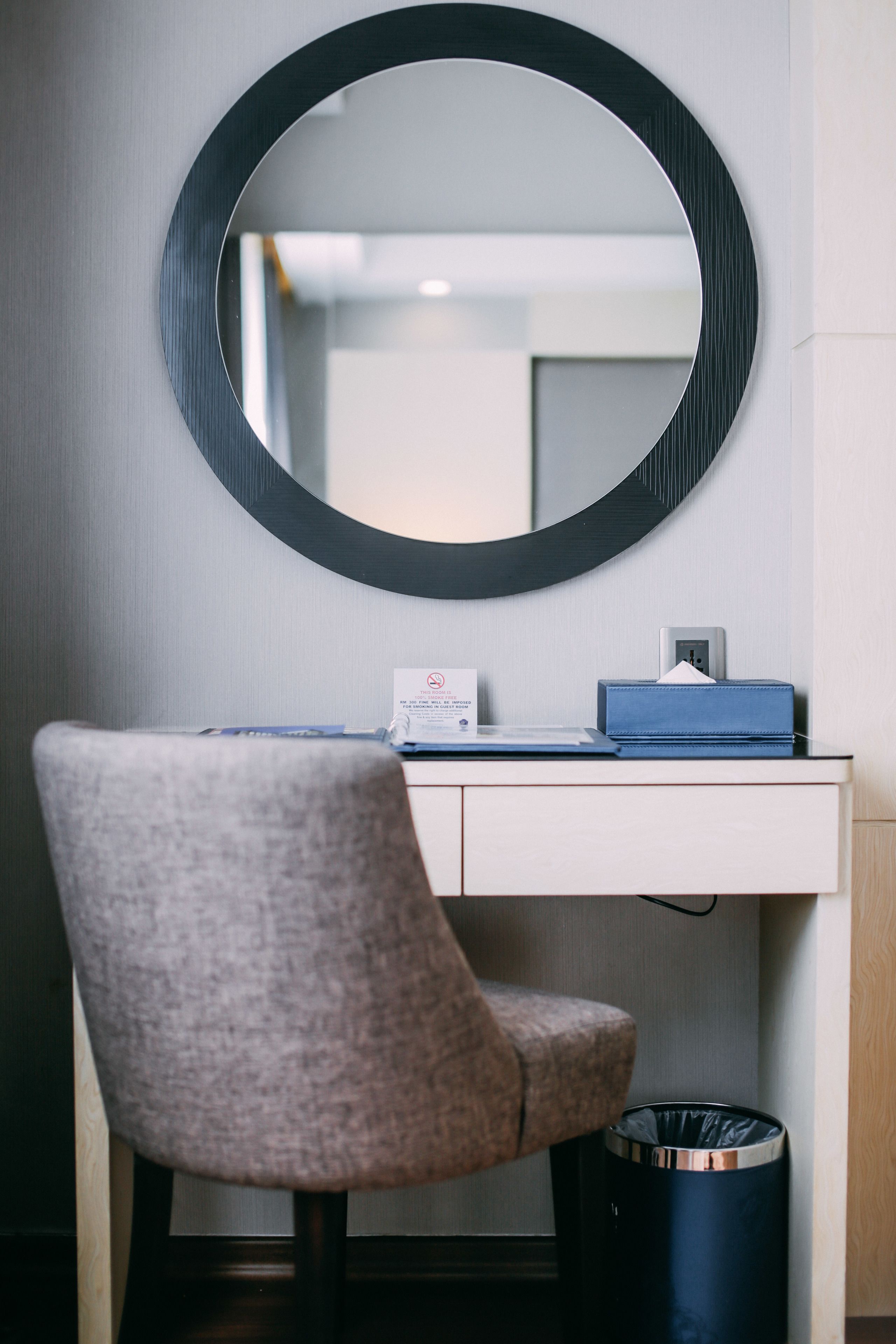 A room with a round mirror on the wall, a desk with a blue box and a card, a grey chair, and a trash bin.