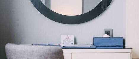 A room with a round mirror on the wall, a desk with a blue box and a card, a grey chair, and a trash bin.