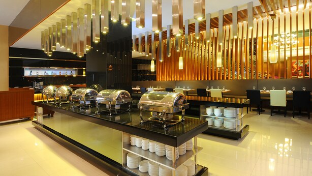 Montaje tipo bufé con varias estaciones de servicio, una barra con platos apilados y un diseño interior moderno con lámparas verticales.