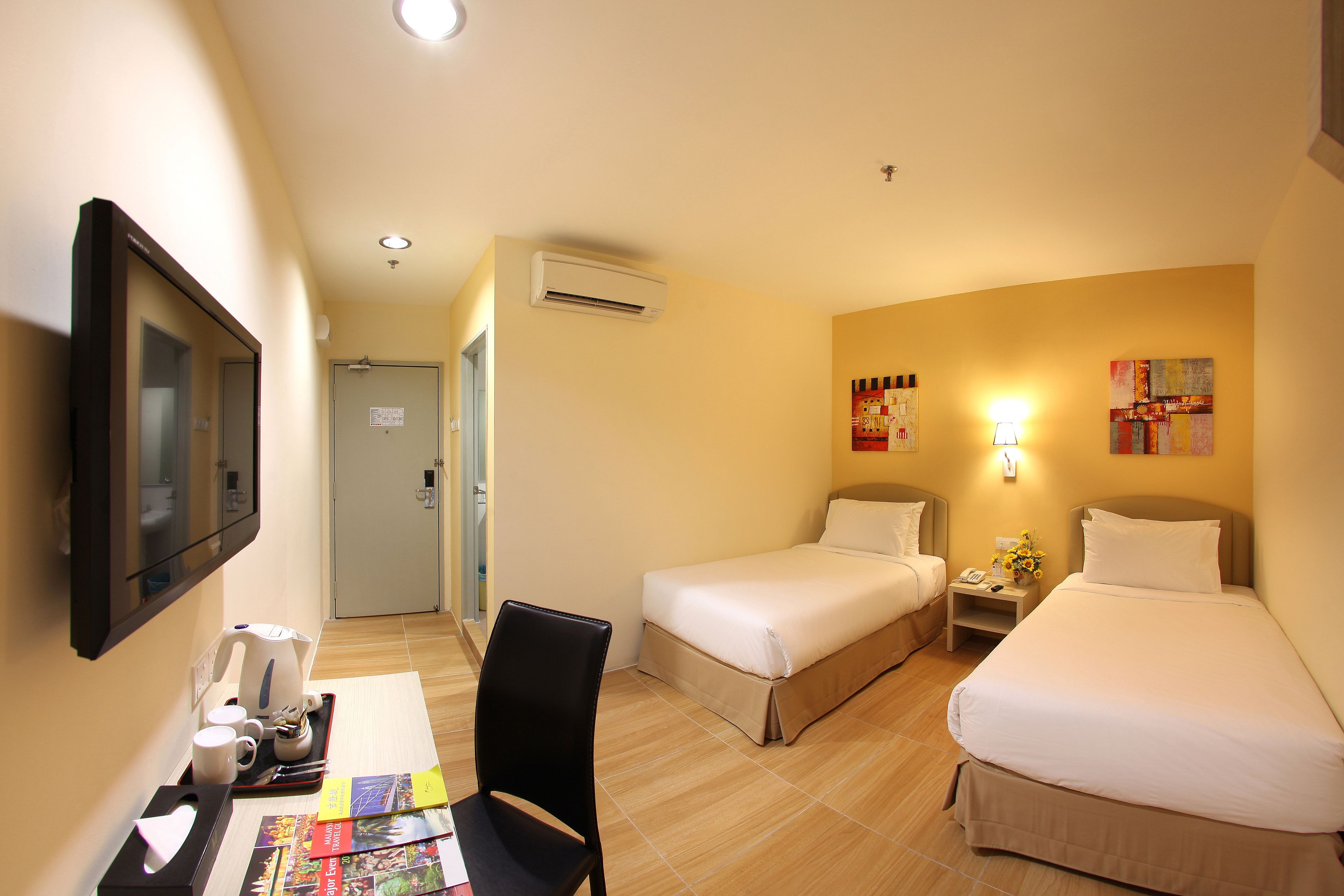 A hotel room with two beds, a desk with a chair, a TV, and a small table with a lamp and flowers.