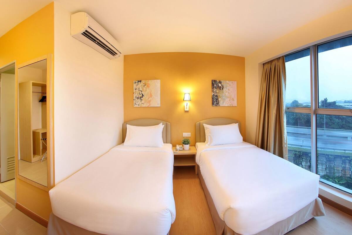 A hotel room with two beds, a bedside table with a lamp, a small plant, and a window with a view of a road.